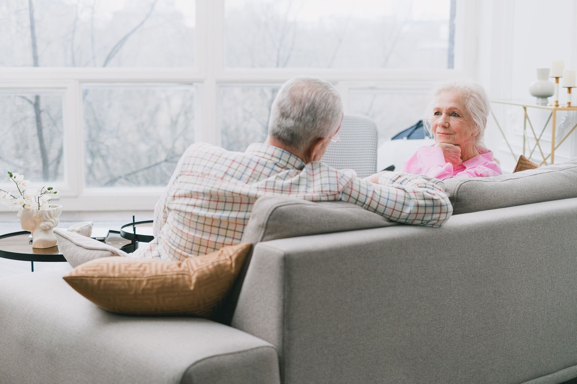 Elderly couple sitting together on a sofa, engaged in a warm conversation inside a well-lit room.